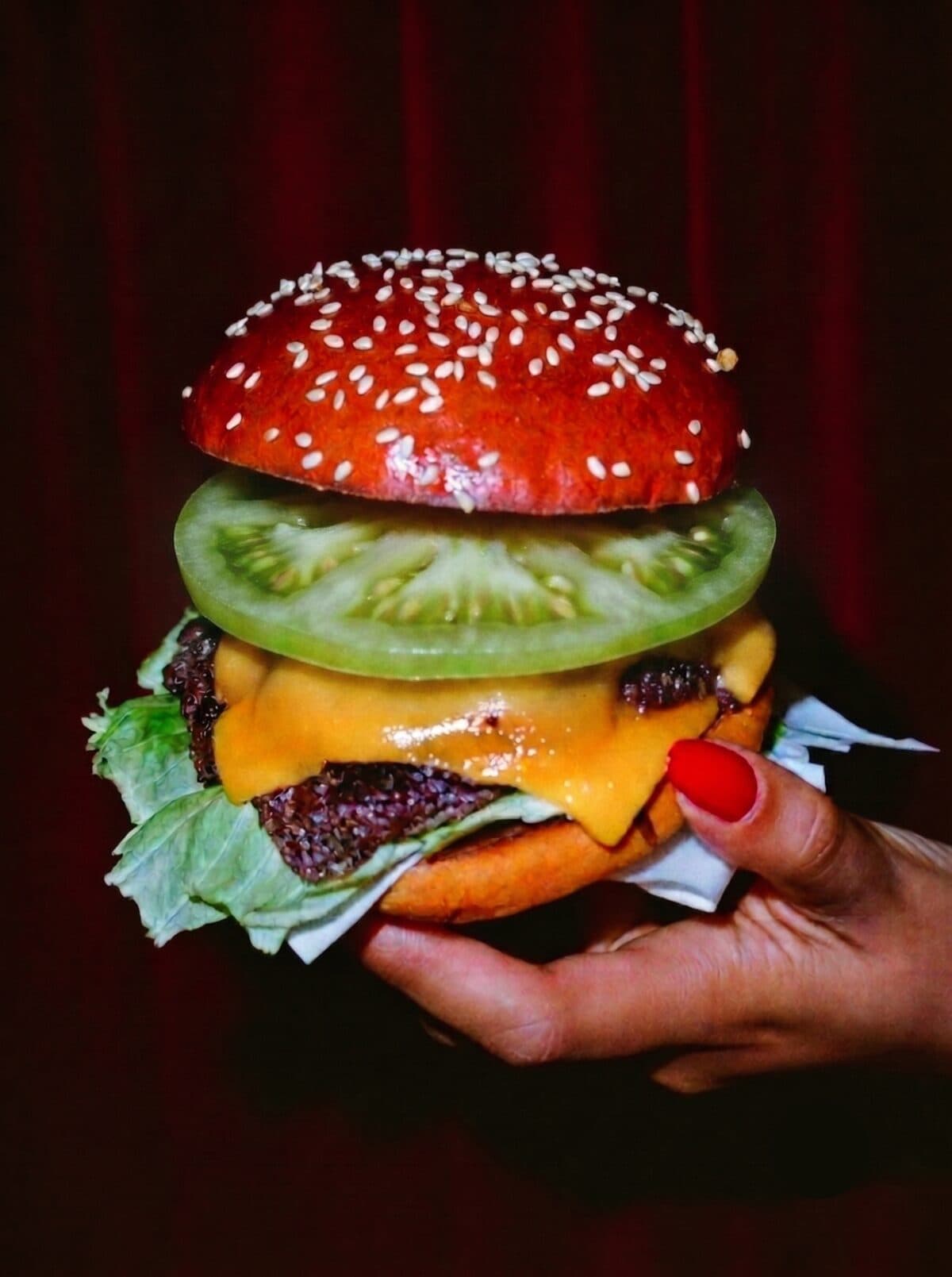 A hand with red nails holding a sesame-seed-bun cheeseburger with a thick slice of green tomato