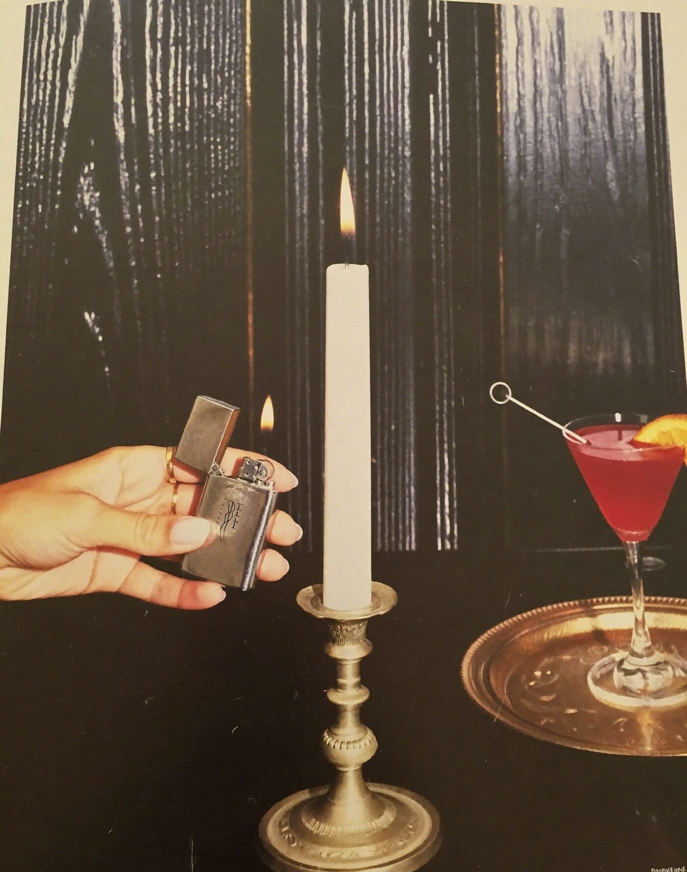 A hand lighting a taper candle with a vintage lighter beside a cocktail on a brass tray
