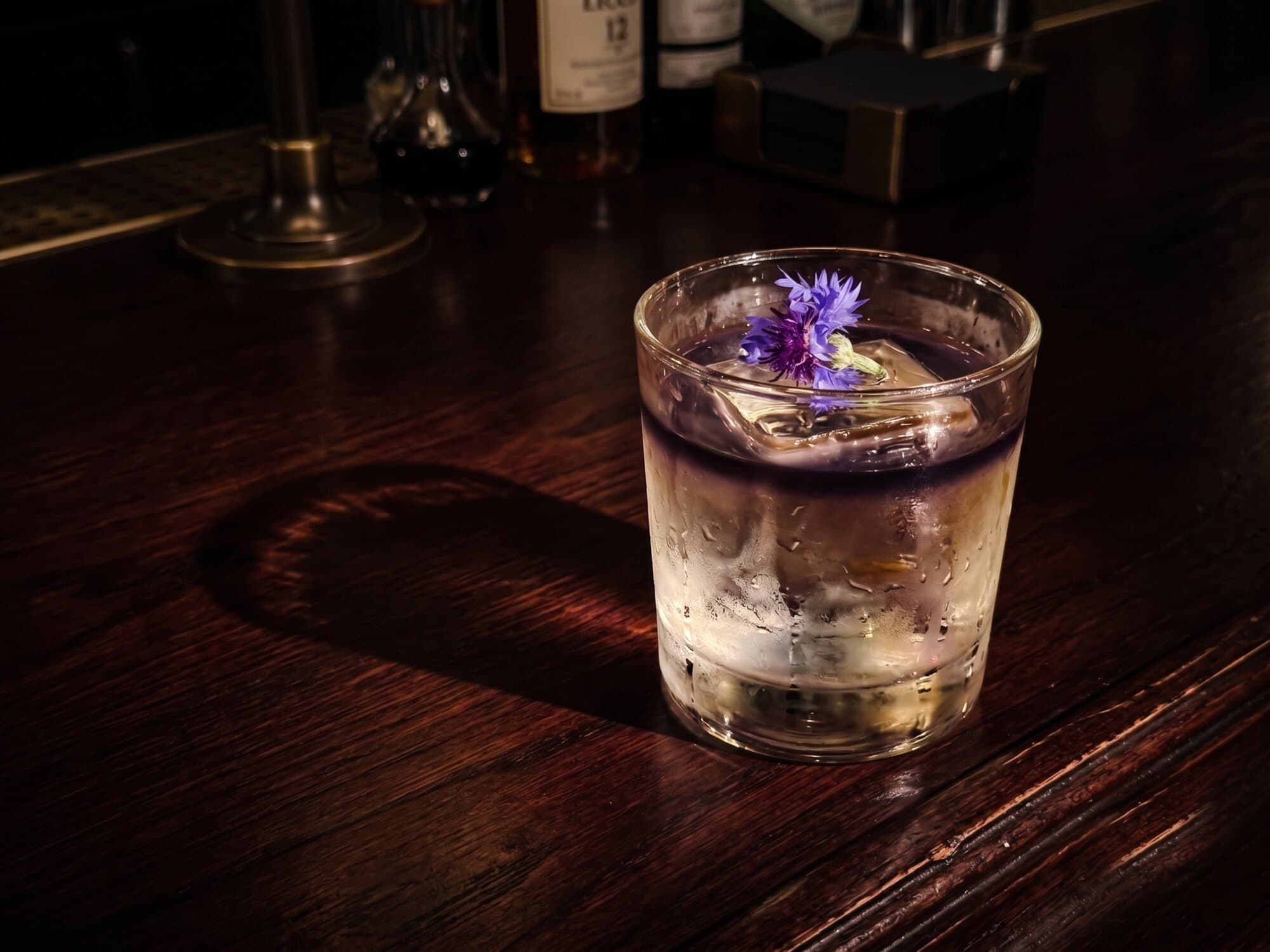 A cocktail over ice with a blue cornflower garnish on a dark wood bar, whiskey bottles behind